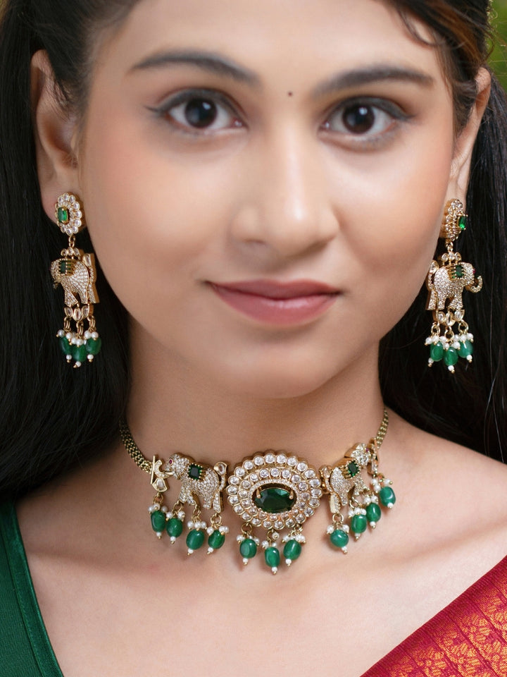 A woman wearing the Aneya Dark Green Choker Necklace Set, featuring ornate gold-toned jewelry with Dark Green and white colored stones and elephant-shaped motifs in the necklace and earrings, poses in traditional attire, her long dark hair partially visible.