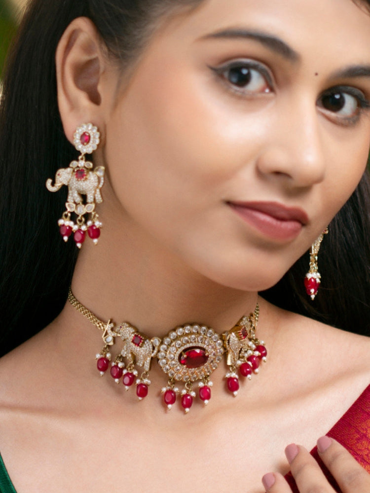 A woman wearing the Aneya Maroon Choker Necklace Set, featuring ornate gold-toned jewelry with red and white colored stones and elephant-shaped motifs in the necklace and earrings, poses in traditional attire, her long dark hair partially visible.