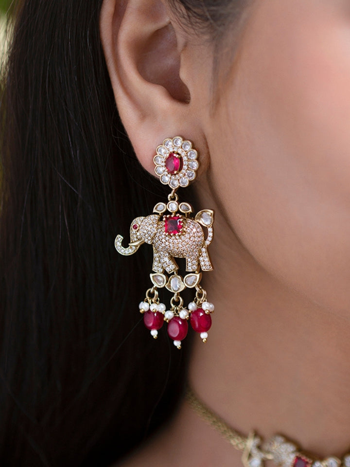 A woman wearing the Aneya Maroon Choker Necklace Set, featuring ornate gold-toned jewelry with red and white colored stones and elephant-shaped motifs in the necklace and earrings, poses in traditional attire, her long dark hair partially visible.