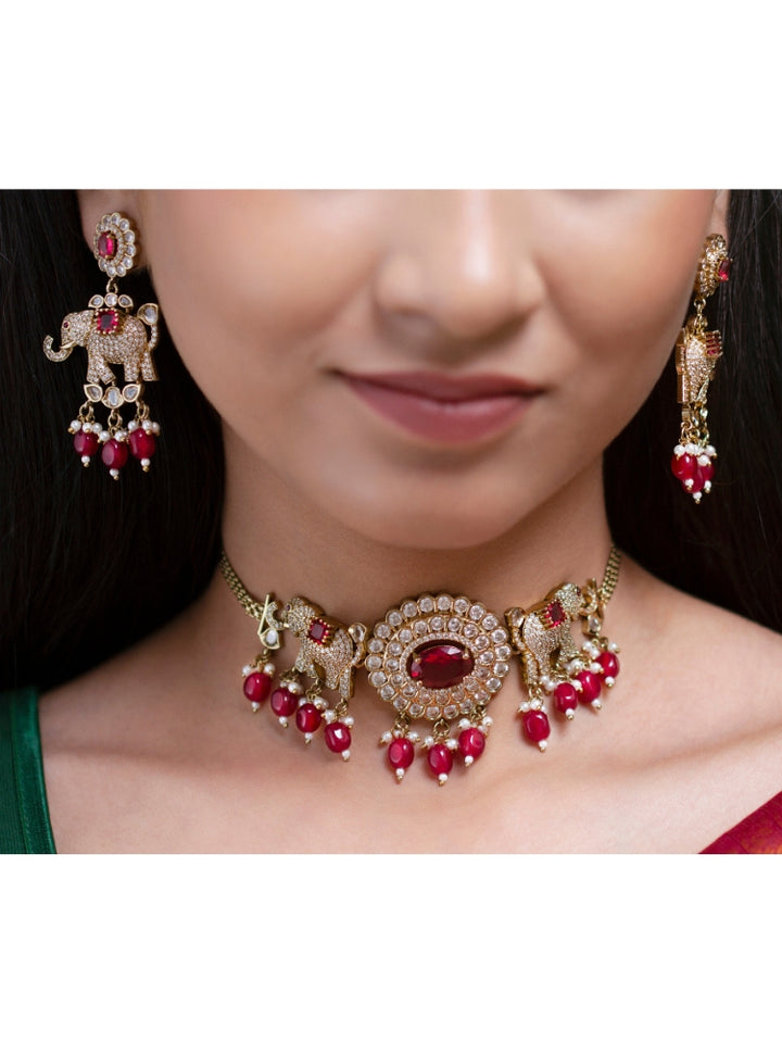 A woman wearing the Aneya Maroon Choker Necklace Set, featuring ornate gold-toned jewelry with red and white colored stones and elephant-shaped motifs in the necklace and earrings, poses in traditional attire, her long dark hair partially visible.
