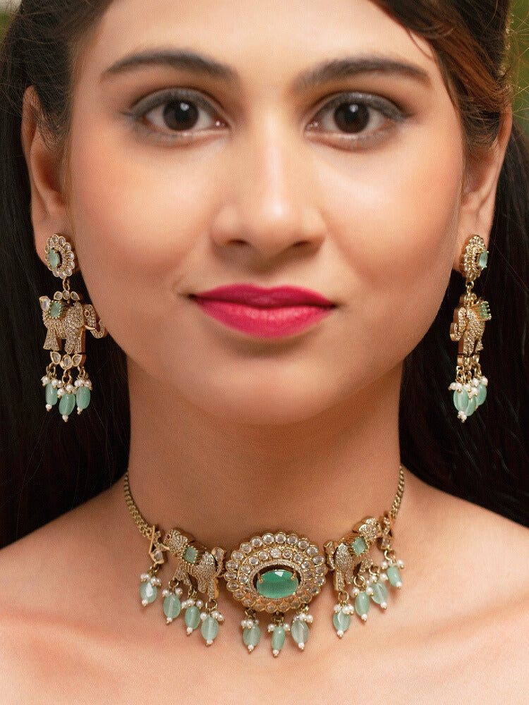 A woman wearing the Aneya Mint Green Choker Necklace Set, featuring ornate gold-toned jewelry with Mint Green and white colored stones and elephant-shaped motifs in the necklace and earrings, poses in traditional attire, her long dark hair partially visible.