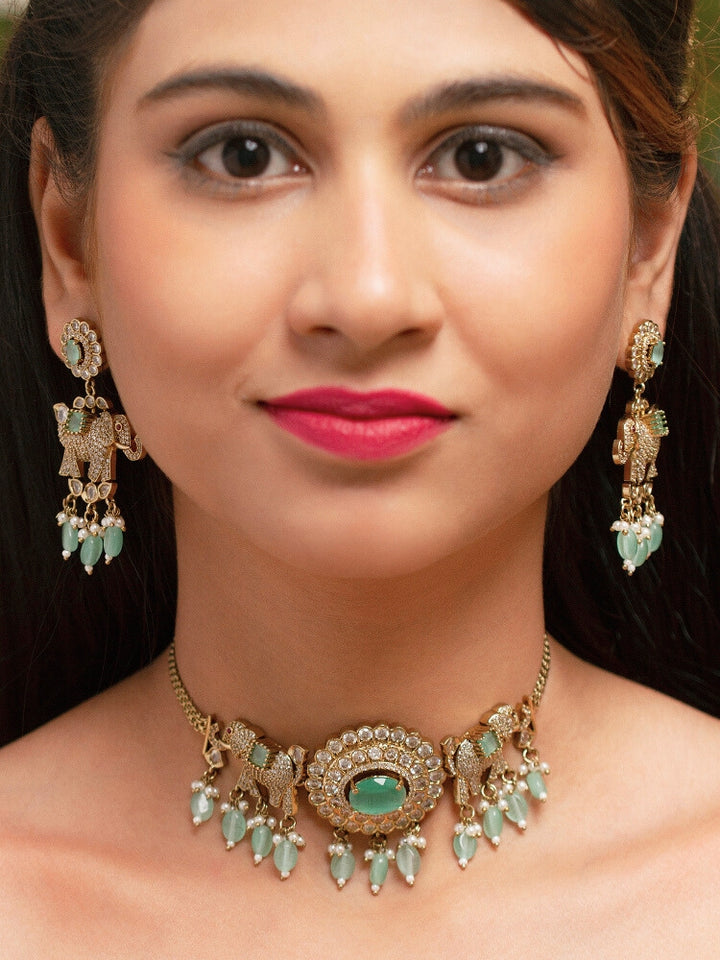 A woman wearing the Aneya Mint Green Choker Necklace Set, featuring ornate gold-toned jewelry with Mint Green and white colored stones and elephant-shaped motifs in the necklace and earrings, poses in traditional attire, her long dark hair partially visible.