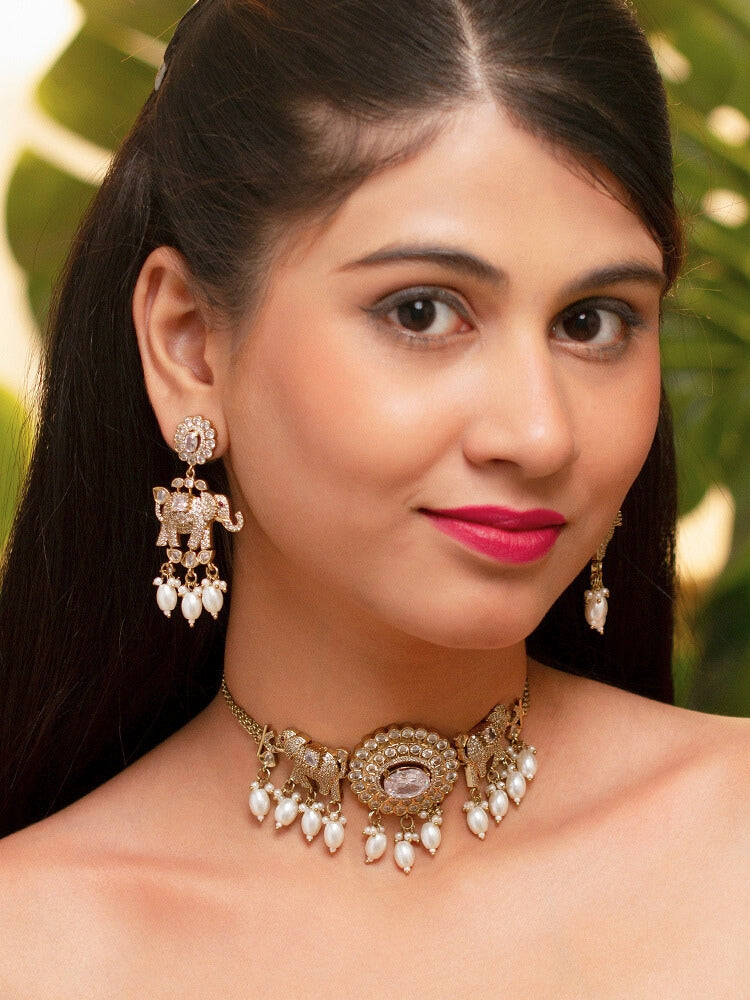 A woman wearing the Aneya White  Choker Necklace Set, featuring ornate gold-toned jewelry with white colored stones and elephant-shaped motifs in the necklace and earrings, poses in traditional attire, her long dark hair partially visible.