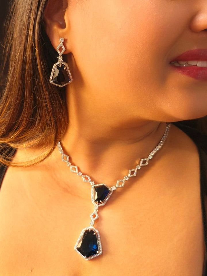 A woman showcases the Angela Blue Necklace Set, featuring matching jewelry with bold Blue stones and sparkling American Diamonds on both drop earrings and a geometric pendant necklace for a striking look.