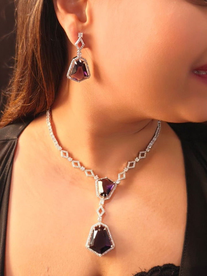 A woman showcases the Angela Purple Necklace Set, featuring matching jewelry with bold Purple stones and sparkling American Diamonds on both drop earrings and a geometric pendant necklace for a striking look.