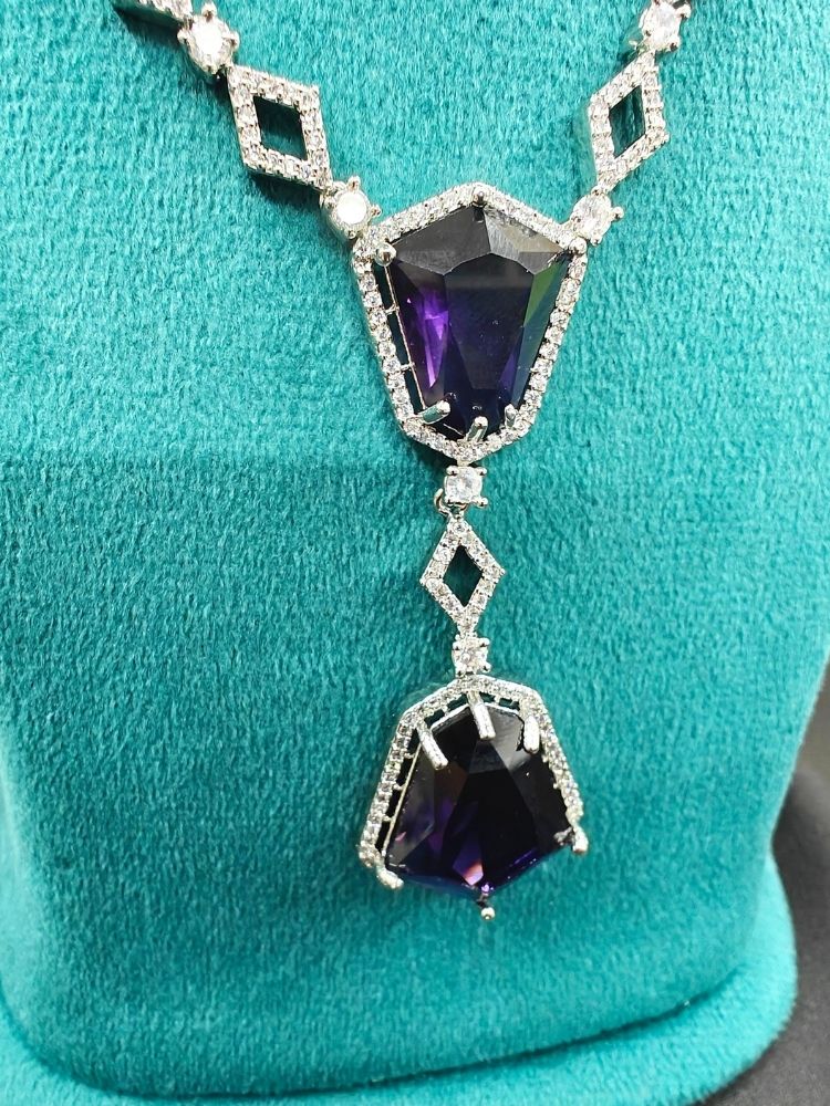 Angela Purple Necklace Set, featuring matching jewelry with bold Purple stones and sparkling American Diamonds on both drop earrings and a geometric pendant necklace for a striking look. Set on a blue dummy. 