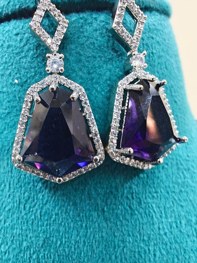 Angela Purple Necklace Set, featuring matching jewelry with bold Purple stones and sparkling American Diamonds on both drop earrings and a geometric pendant necklace for a striking look. Set on a blue dummy. 