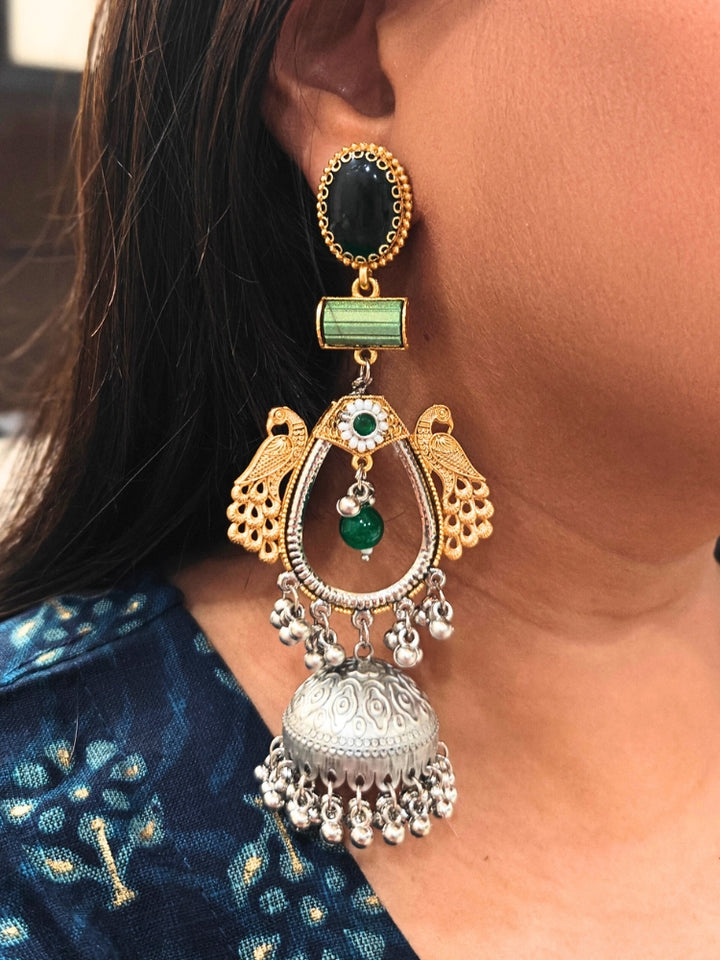 A close-up of a woman’s ear adorned with the Anvesha Green Earring, featuring gold peacocks, a green stone, and a large silver jhumka accented with brass pearls, intricate designs, and small hanging beads. Long Oxidized earrings, german silver earrings, navratri earrings. 