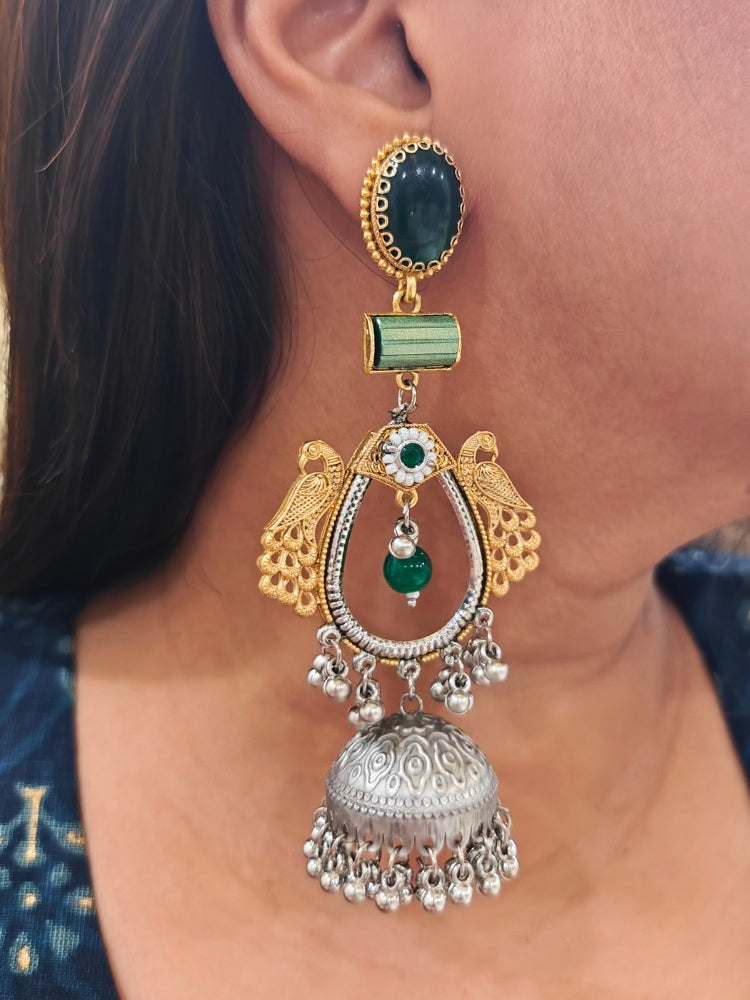 A close-up of a woman’s ear adorned with the Anvesha Green Earring, featuring gold peacocks, a green stone, and a large silver jhumka accented with brass pearls, intricate designs, and small hanging beads. Long Oxidized earrings, german silver earrings, navratri earrings. 