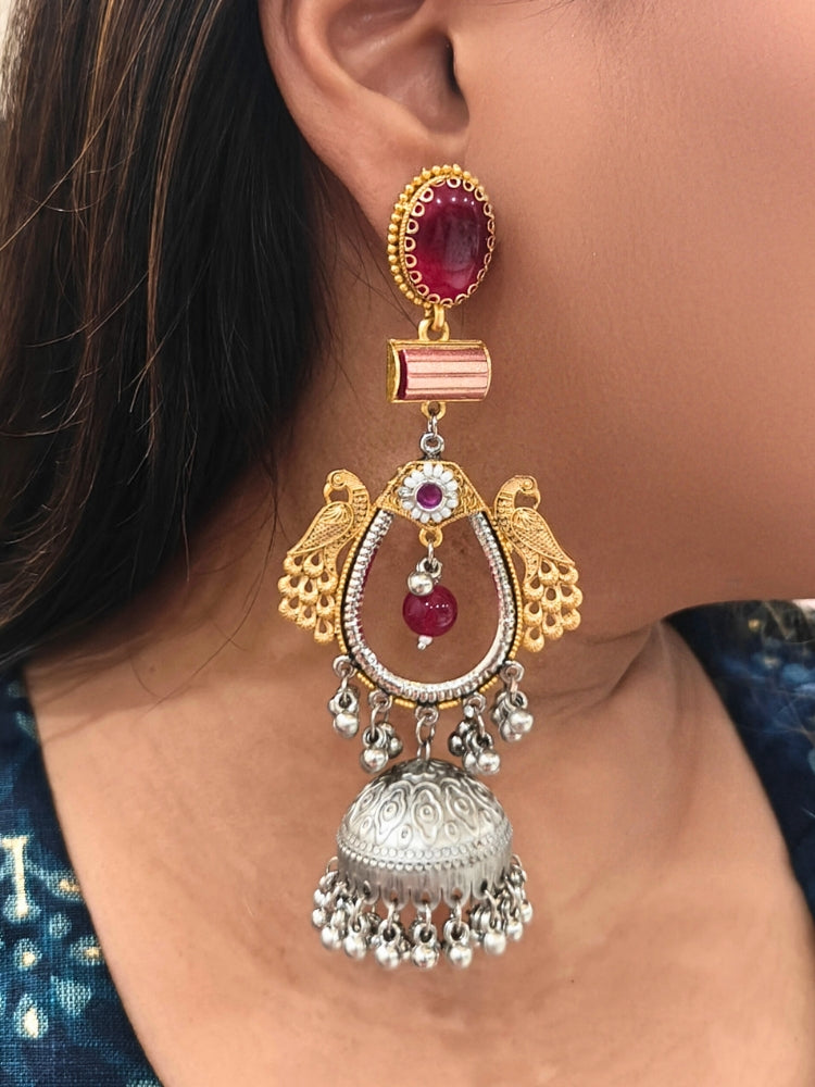 A close-up of a woman’s ear adorned with the Anvesha Red Earring, featuring gold peacocks, a red stone, and a large silver jhumka accented with brass pearls, intricate designs, and small hanging beads. Oxidized earrings, german silver earrings, navratri earrings. 