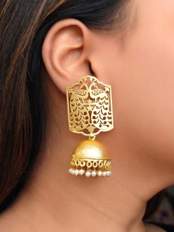 A close-up of a womans ear wearing the Anvita Earrings—gold with intricate filigree, featuring a peacock above a dome-shaped jhumka accented by hanging pearls—perfect to complement your wedding ceremony outfit.