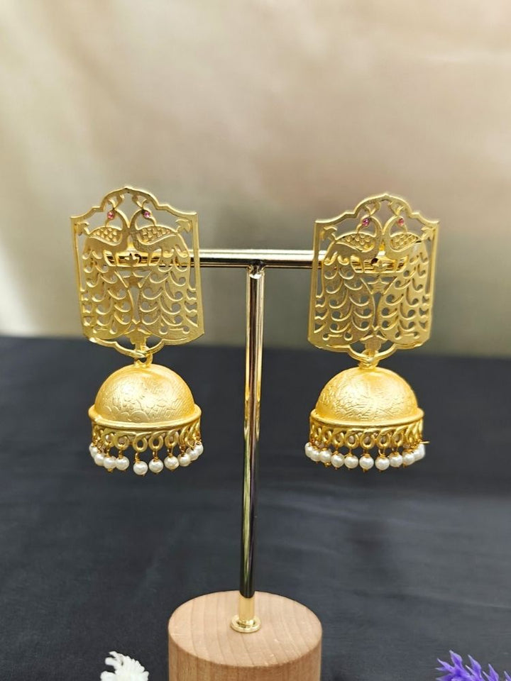 Anvita Earrings—gold with intricate filigree, featuring a peacock above a dome-shaped jhumka accented by hanging pearls—perfect to complement your wedding ceremony outfit, shown on an earrings holder with black background. 