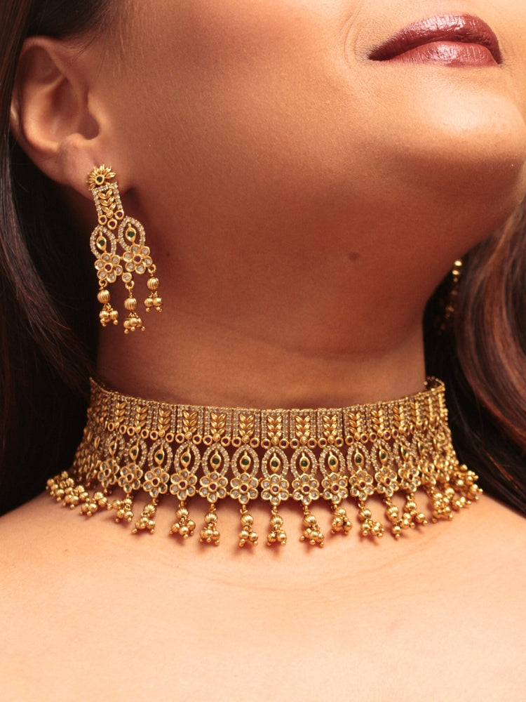 A woman models the Ashira Choker Necklace Set, featuring an ornate gold choker with intricate detailing with red and green colored stones and dangling beads, paired with matching earrings. She wears glossy brown lipstick and her hair falls softly to one side.