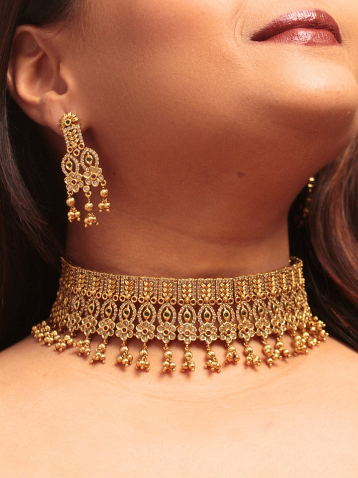A woman models the Ashira Choker Necklace Set, featuring an ornate gold choker with intricate detailing with red and green colored stones and dangling beads, paired with matching earrings. She wears glossy brown lipstick and her hair falls softly to one side.