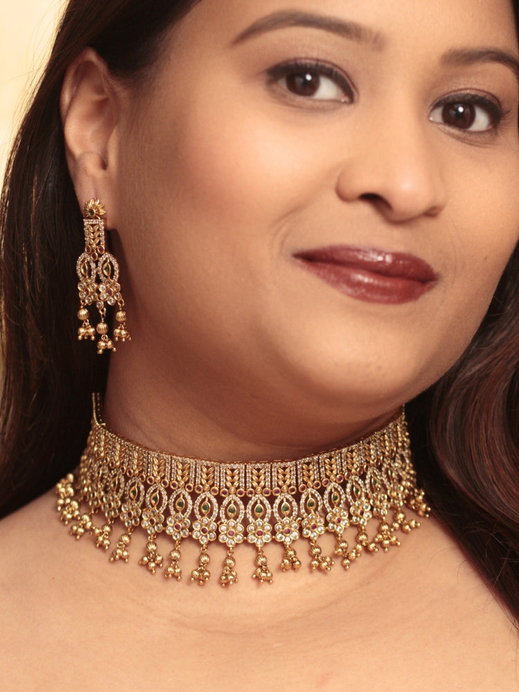 A woman models the Ashira Choker Necklace Set, featuring an ornate gold choker with intricate detailing with red and green colored stones and dangling beads, paired with matching earrings. She wears glossy brown lipstick and her hair falls softly to one side.