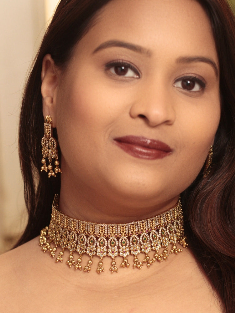 A woman models the Ashira Choker Necklace Set, featuring an ornate gold choker with intricate detailing with red and green colored stones and dangling beads, paired with matching earrings. She wears glossy brown lipstick and her hair falls softly to one side.