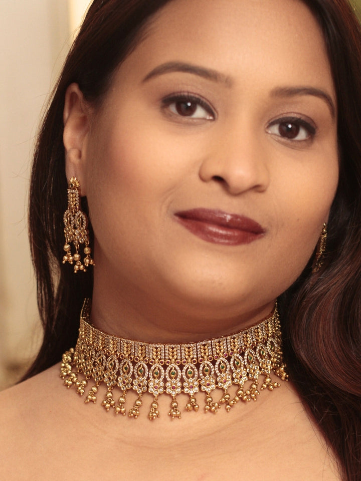 A woman models the Ashira Choker Necklace Set, featuring an ornate gold choker with intricate detailing with red and green colored stones and dangling beads, paired with matching earrings. She wears glossy brown lipstick and her hair falls softly to one side.