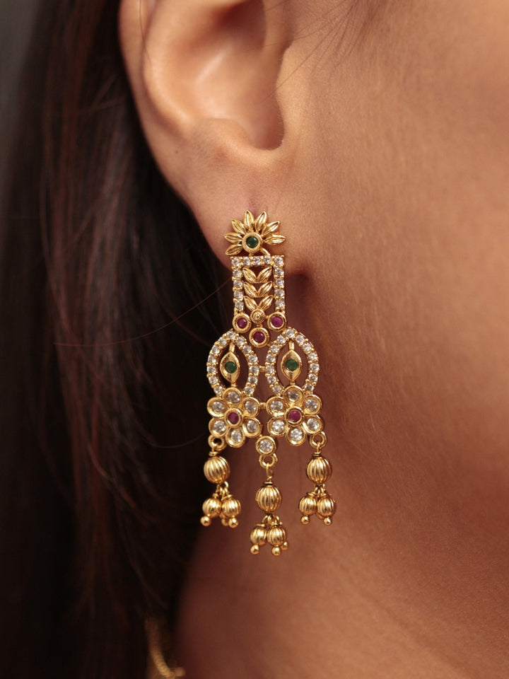 A woman models the Ashira Choker Necklace Set, featuring an ornate gold choker with intricate detailing with red and green colored stones and dangling beads, paired with matching earrings. She wears glossy brown lipstick and her hair falls softly to one side.