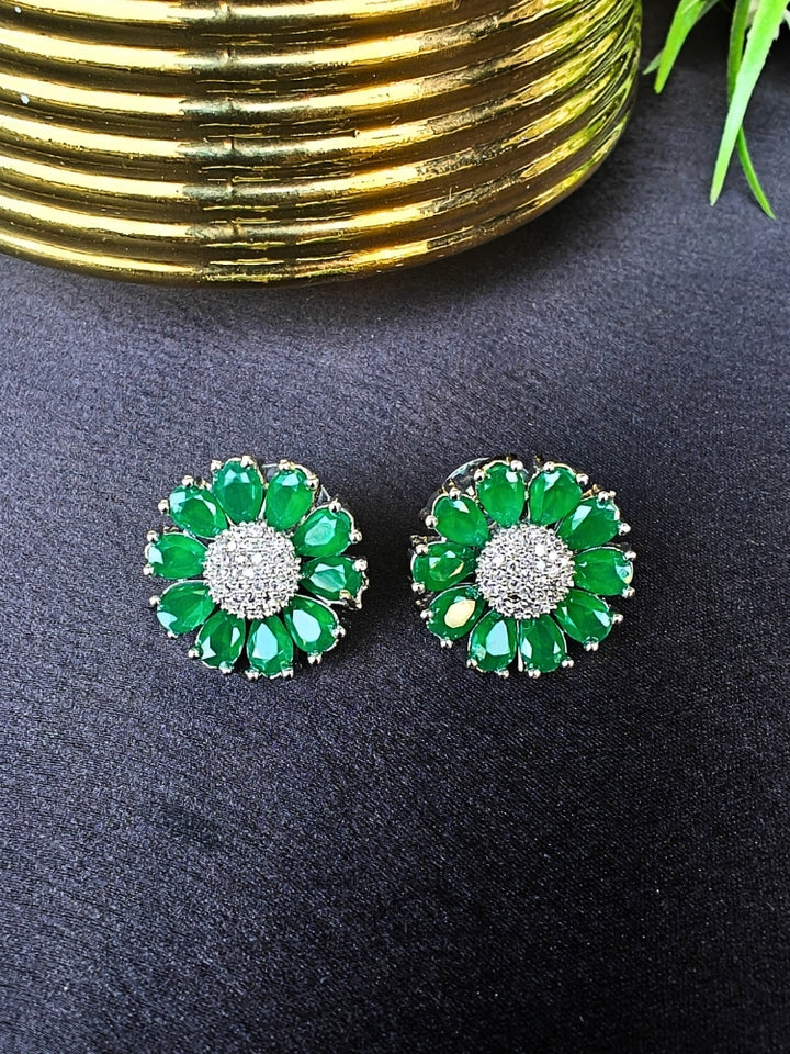 The Aspen Green stud Earrings are round, flower-shaped with green petals and a central cluster of american diamonds. Shown on a black surface beside a gold-striped object and a plant.