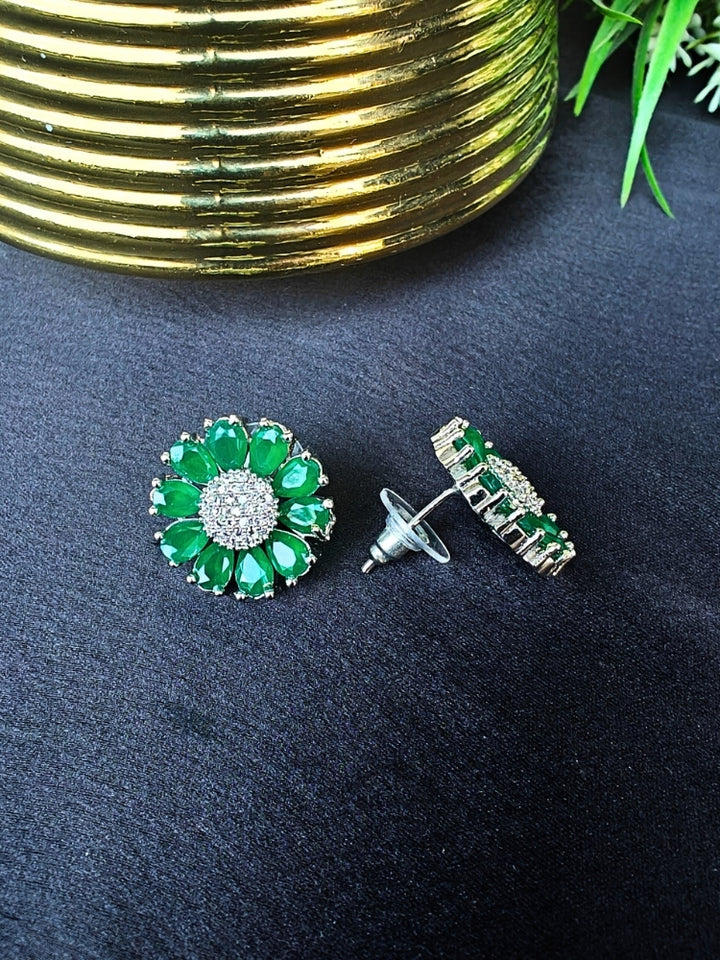 The Aspen Green stud Earrings are round, flower-shaped with green petals and a central cluster of american diamonds. Shown on a black surface beside a gold-striped object and a plant.