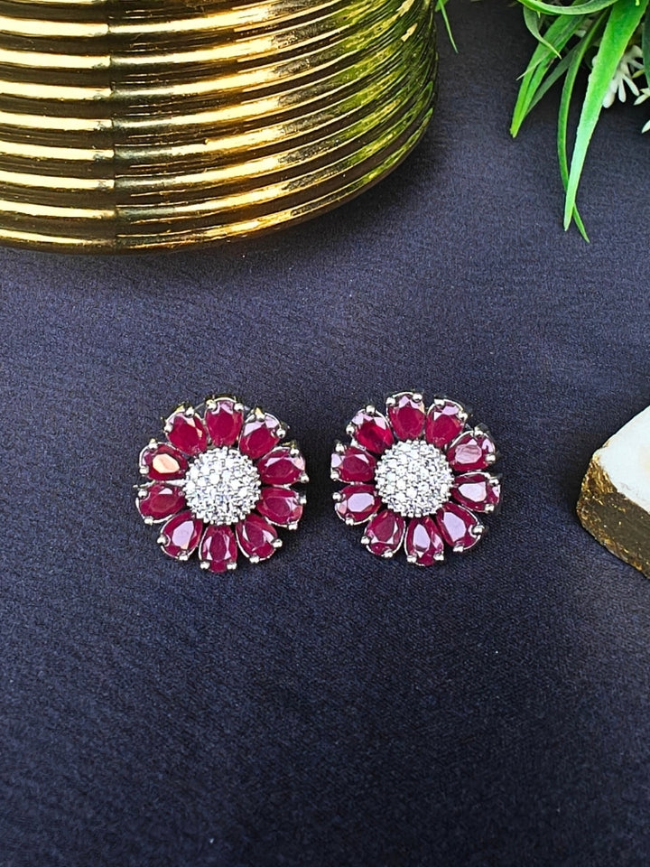 The Aspen Pink stud Earrings are round, flower-shaped with Pink petals and a central cluster of american diamonds. Shown on a black surface beside a gold-striped object and a plant.