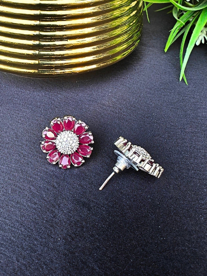 The Aspen Pink stud Earrings are round, flower-shaped with Pink petals and a central cluster of american diamonds. Shown on a black surface beside a gold-striped object and a plant.