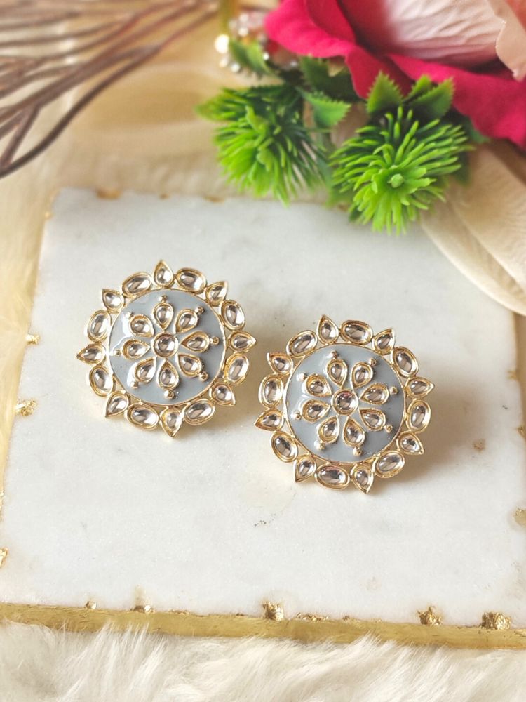 The Audrey Grey stud Earrings are round, gold-toned Kundan earrings, shown on a white marble slab with green leaves and red flowers in the background.