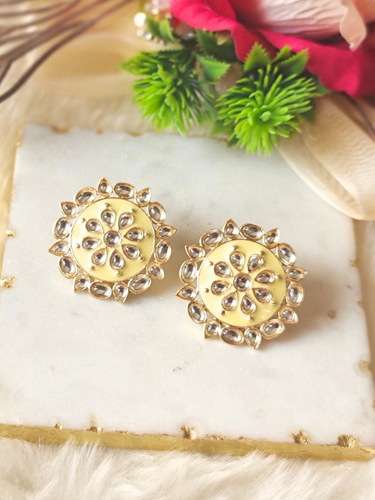The Audrey Mint Yellow stud Earrings are round, gold-toned Kundan earrings, shown on a white marble slab with green leaves and red flowers in the background.