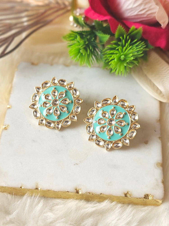 The Audrey Sea Green stud Earrings are round, gold-toned Kundan earrings, shown on a white marble slab with green leaves and red flowers in the background.