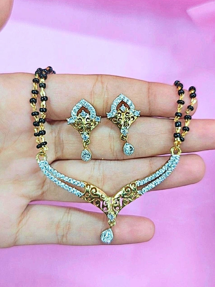 A hand holds the Avantika Mangalsutra—a gold necklace with black mangalsutra beads and matching earrings, pendant featuring sparkling American Diamonds—set against a soft pink background, perfect for your wedding ceremony dress. Diamond mangalsutra. Modern mangalsutra. 