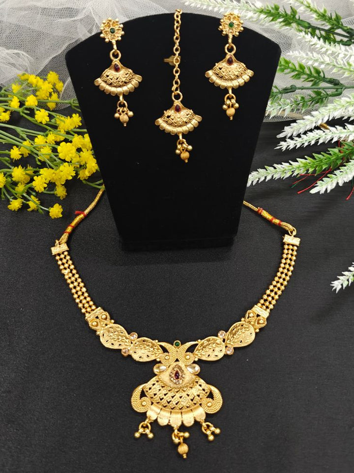 The Avyan Necklace Set is on display, featuring an ornate wedding gold necklace with a central pendant and matching earrings adorned with red gemstone accents. Intricate designs add elegance, while yellow flowers and greenery decorate the background.