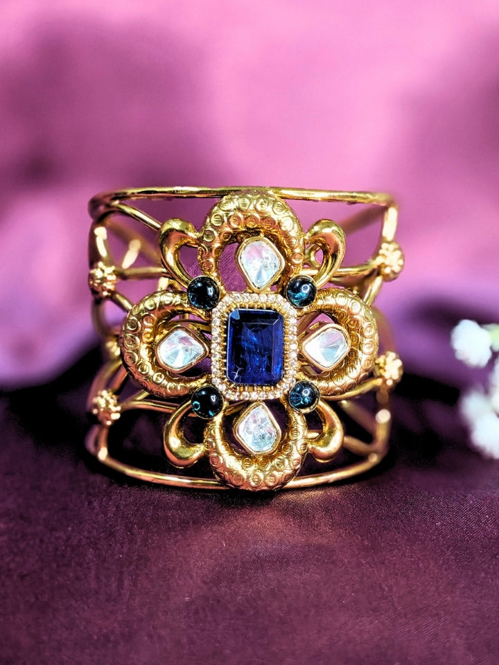 The Aziza Navy Blue Bracelet (Adjustable) showcases an intricate floral design with a large Navy Blue center stone, kundan and smaller Navy Blue colored stones, set against a rich purple background—an ideal choice for wedding jewelry.