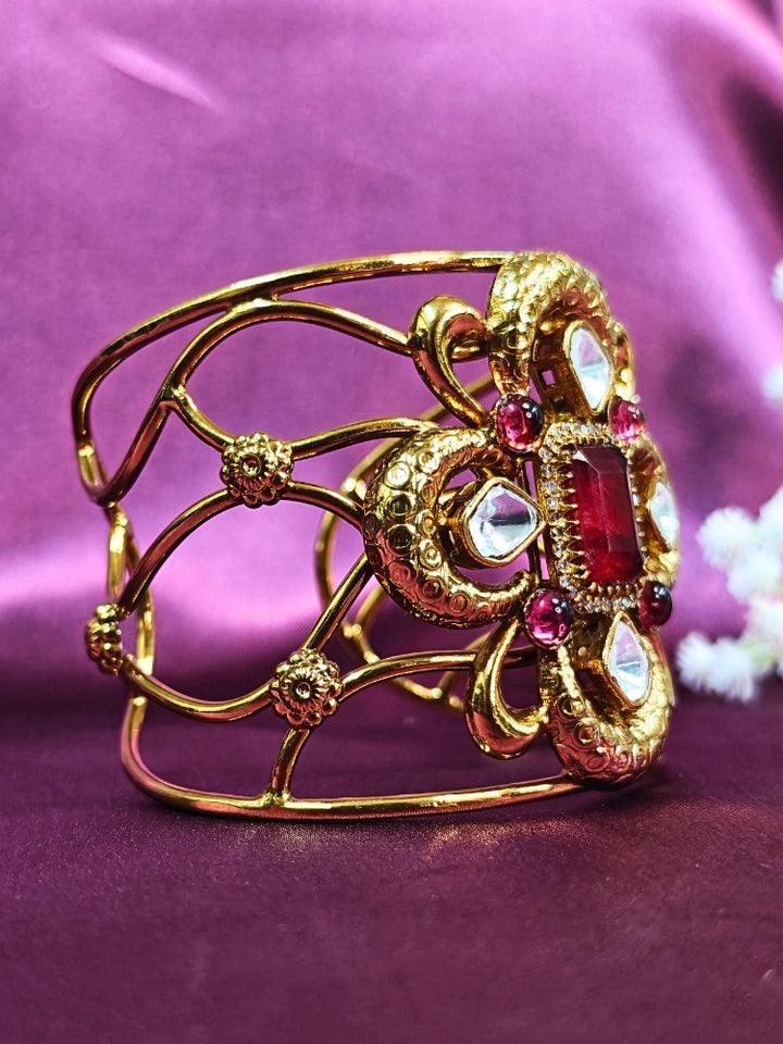 The Aziza Red Bracelet (Adjustable) showcases an intricate floral design with a large red center stone, kundan and smaller red colored stones, set against a rich purple background—an ideal choice for wedding jewelry.
