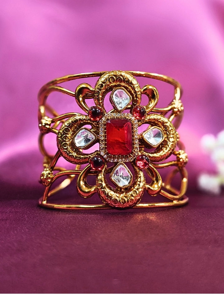 The Aziza Red Bracelet (Adjustable) showcases an intricate floral design with a large red center stone, kundan and smaller red colored stones, set against a rich purple background—an ideal choice for wedding jewelry.