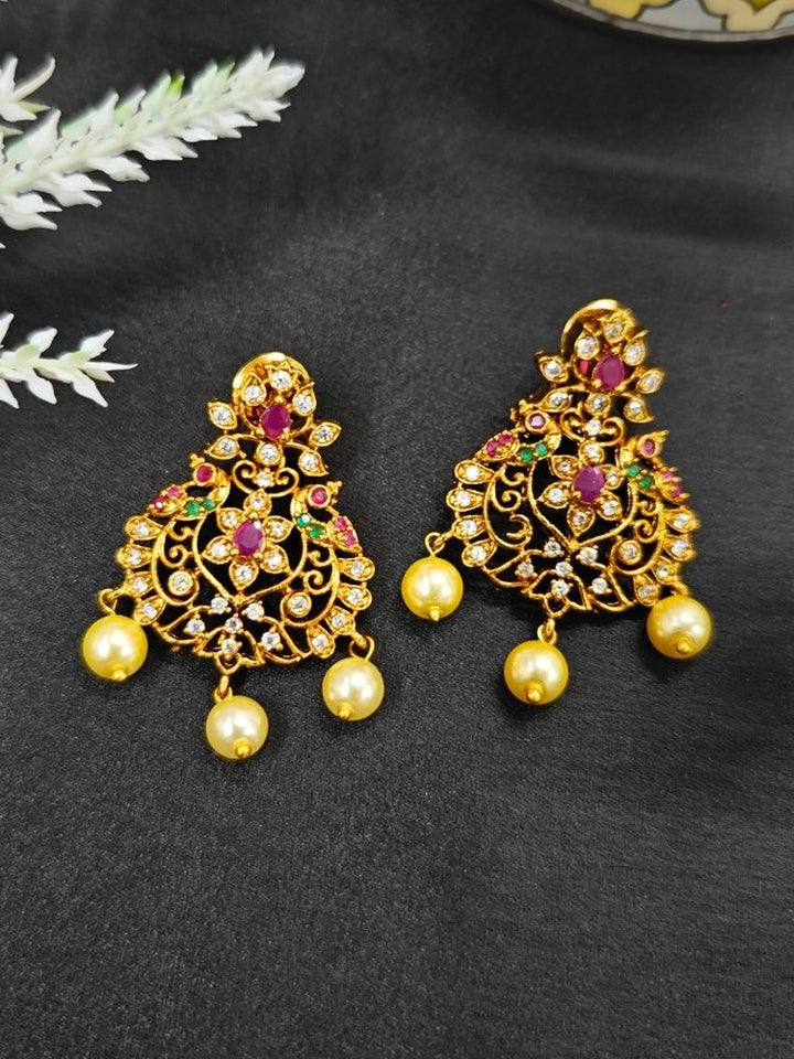 The Bhakti Earrings are ornate gold earrings with intricate filigree, studded with red and white stones and American Diamonds, each featuring three dangling round yellow pearls, elegantly displayed on a black fabric background.