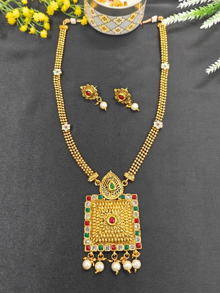 The Bharti Necklace Set features a gold necklace with intricate filigree, a large square pendant embellished with green and red stones, pearl drops, and matching earrings, showcased on a black background with yellow flowers in the corner.