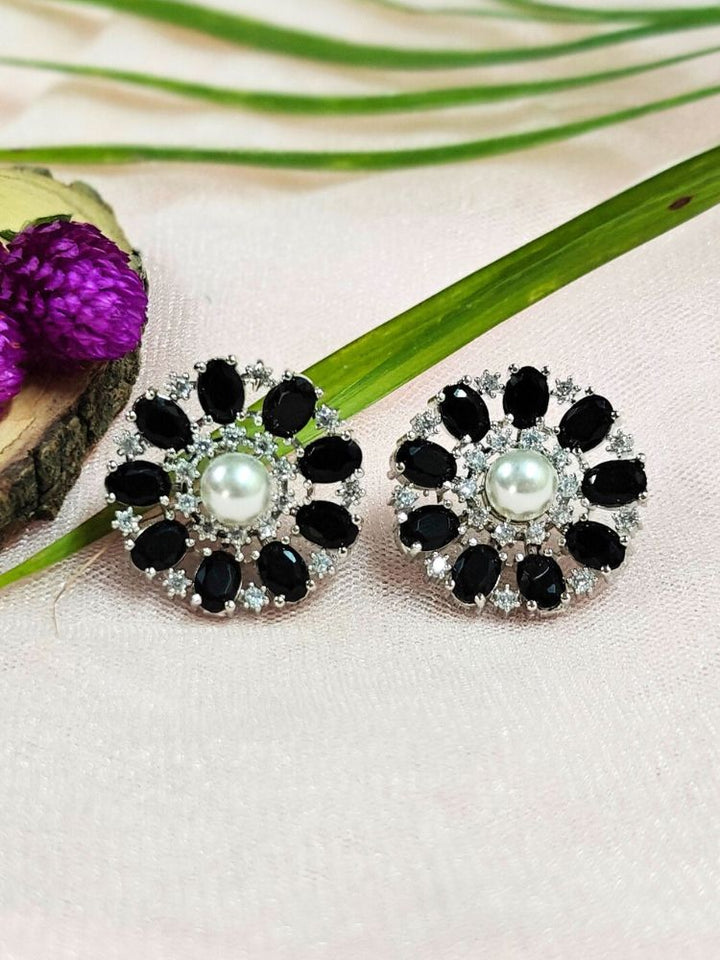 The Black Blossom Earrings showcase a central white pearl encircled by alternating black stones and American diamonds, elegantly displayed on soft pink fabric with green leaves and a purple flower.