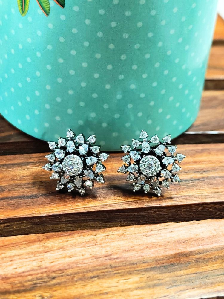 The Black Dahlia stud Earrings are chic, flower-shaped Black stud earrings adorned with American diamonds, elegantly displayed on a wooden surface against a green polka-dotted background.