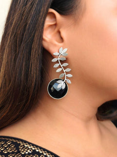 A close-up of a womans ear wearing Harriet Black Earrings—large brass with a leafy branch design and a round, translucent Black stone at the bottom. Her dark hair and part of a pink patterned garment are also visible. Casual wear earrings. Festive celebration look enhanced with artificial Oxidised Earrings.
