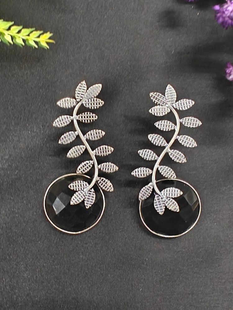 The Harriet Black Earrings feature oxidized polish, leaf-shaped designs on top, and large a large Black round stone at the bottom, displayed on black fabric with green and purple flowers and a patterned ceramic pot in the background.