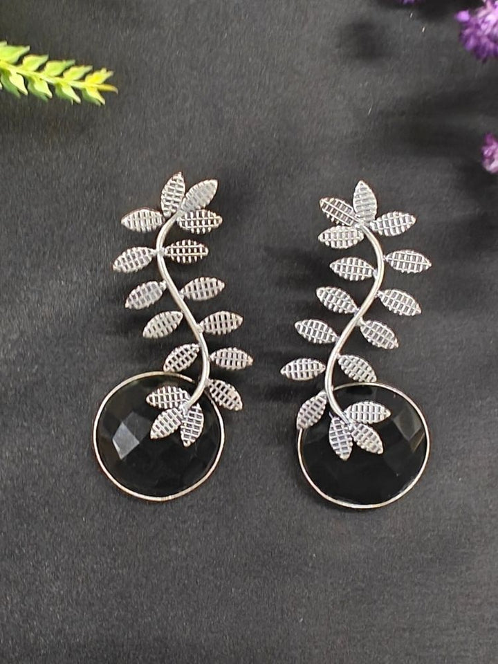The Harriet Black Earrings feature oxidized polish, leaf-shaped designs on top, and large a large Black round stone at the bottom, displayed on black fabric with green and purple flowers and a patterned ceramic pot in the background.