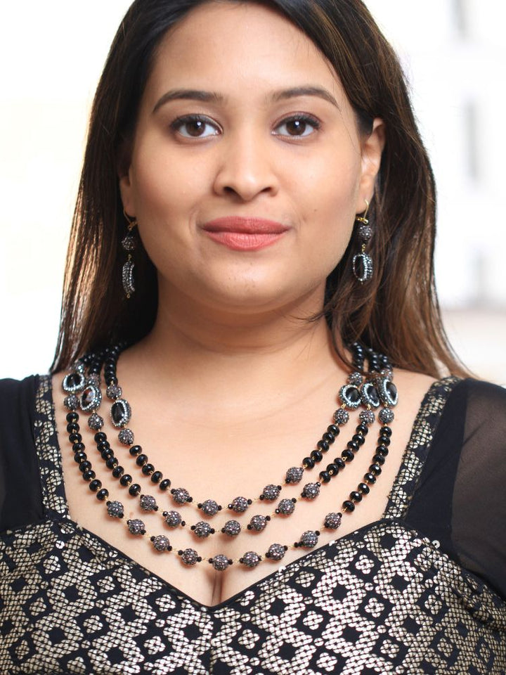 A woman with straight brown hair in Indo-Western attire wears the Black Nebula Long Necklace Set—featuring a patterned black and gold outfit, sparkling earrings, and a layered black beaded necklace—looking neutrally at the camera.
