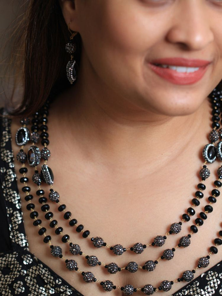 A woman with straight brown hair in Indo-Western attire wears the Black Nebula Long Necklace Set—featuring a patterned black and gold outfit, sparkling earrings, and a layered black beaded necklace—looking neutrally at the camera.