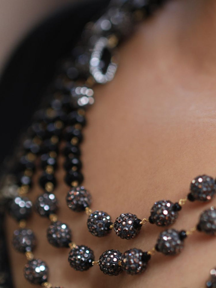 A woman with straight brown hair in Indo-Western attire wears the Black Nebula Long Necklace Set—featuring a patterned black and gold outfit, sparkling earrings, and a layered black beaded necklace—looking neutrally at the camera.