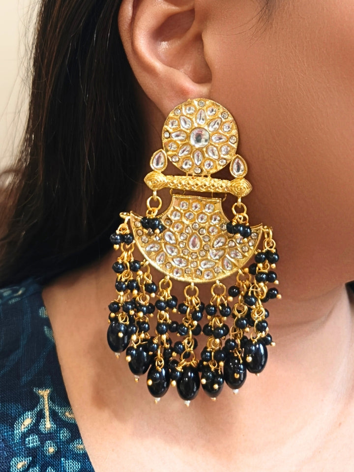 A close-up of a person wearing Black Neha Long Earrings—ornate Kundan designs with American diamonds and dangling black beads—against a blue patterned garment, creating the perfect statement jewelry for any occasion.