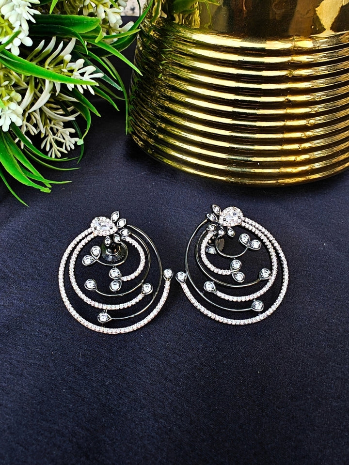 The Black & Pink Maria Earrings feature sparkling circular American Diamonds with intricate spiral designs and clear stones, beautifully displayed on a black surface beside gold decor and green foliage. American Diamond Earrings