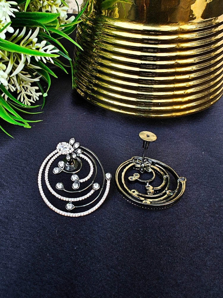 The Black & Pink Maria Earrings feature sparkling circular American Diamonds with intricate spiral designs and clear stones, beautifully displayed on a black surface beside gold decor and green foliage. American Diamond Earrings