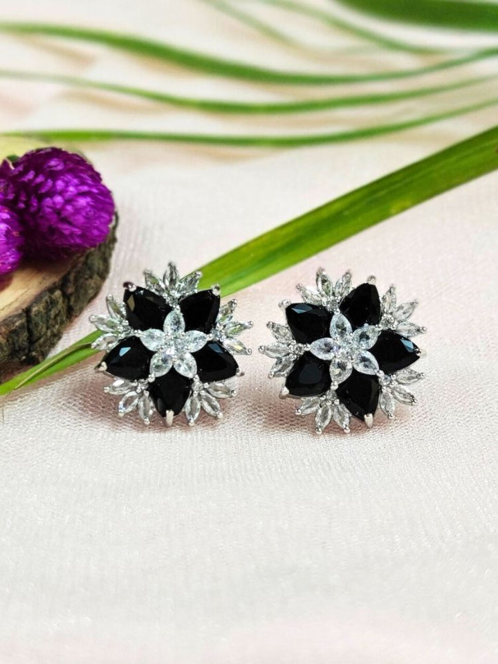 The Black Snowflake Earrings showcase Black colored stones along with American Diamonds in a starburst design, perched on a soft pink fabric with green leaves and a Purple flower.