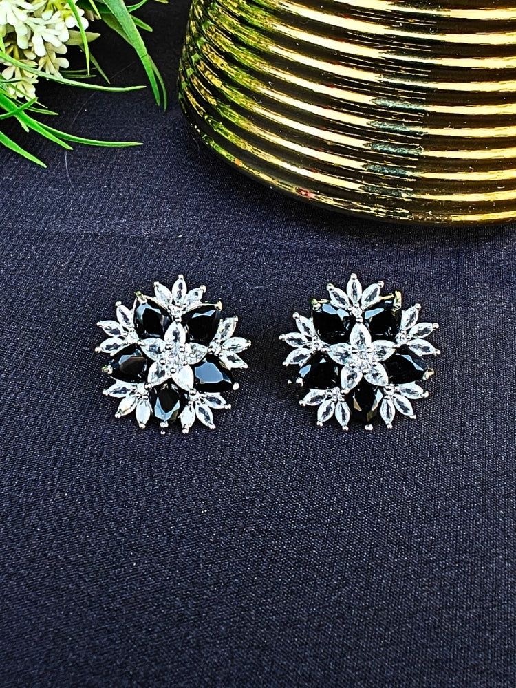 The Black Snowflake Earrings showcase Black colored stones along with American Diamonds in a starburst design, perched on a Black Surface with green leaves.