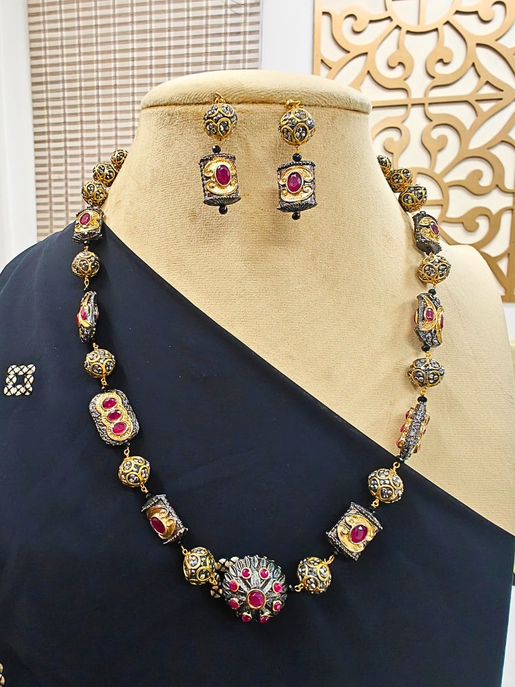 The Black Trisha Necklace Set is a decorative statement accessory with gold and black beads, square accents, and ruby and gold foil work, beautifully displayed on a mannequin in black attire against ornate wall designs.